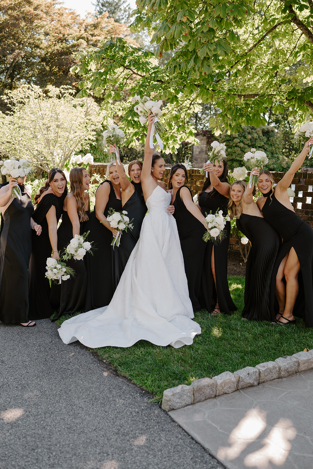 Bride holding her bouquet high as she celebrates with her cheering bridesmaids in black gowns.